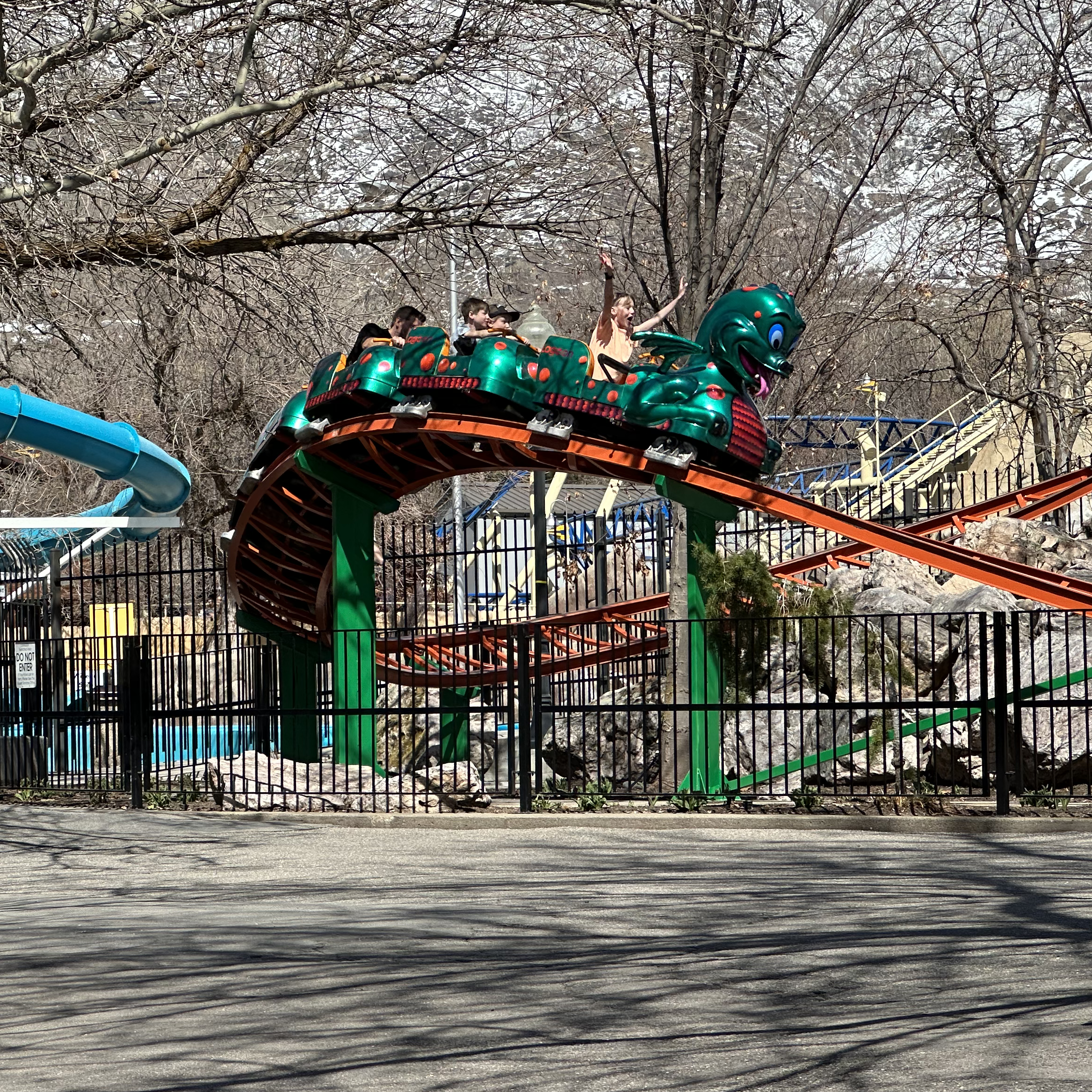 The image shows the Puff the Little Fire Dragon rollercoaster at Lagoon Amusement Park. The rollercoaster features a green dragon-themed train with several cars, each designed to look like a segment of the dragon's body. The train is on a curved section of the track, elevated above the ground and supported by green pillars. In the background, there are leafless trees and parts of other amusement park attractions, including a blue water slide and various structures. The scene is set in an outdoor amusement park, and the rollercoaster appears to be in motion, with riders visible in the cars.