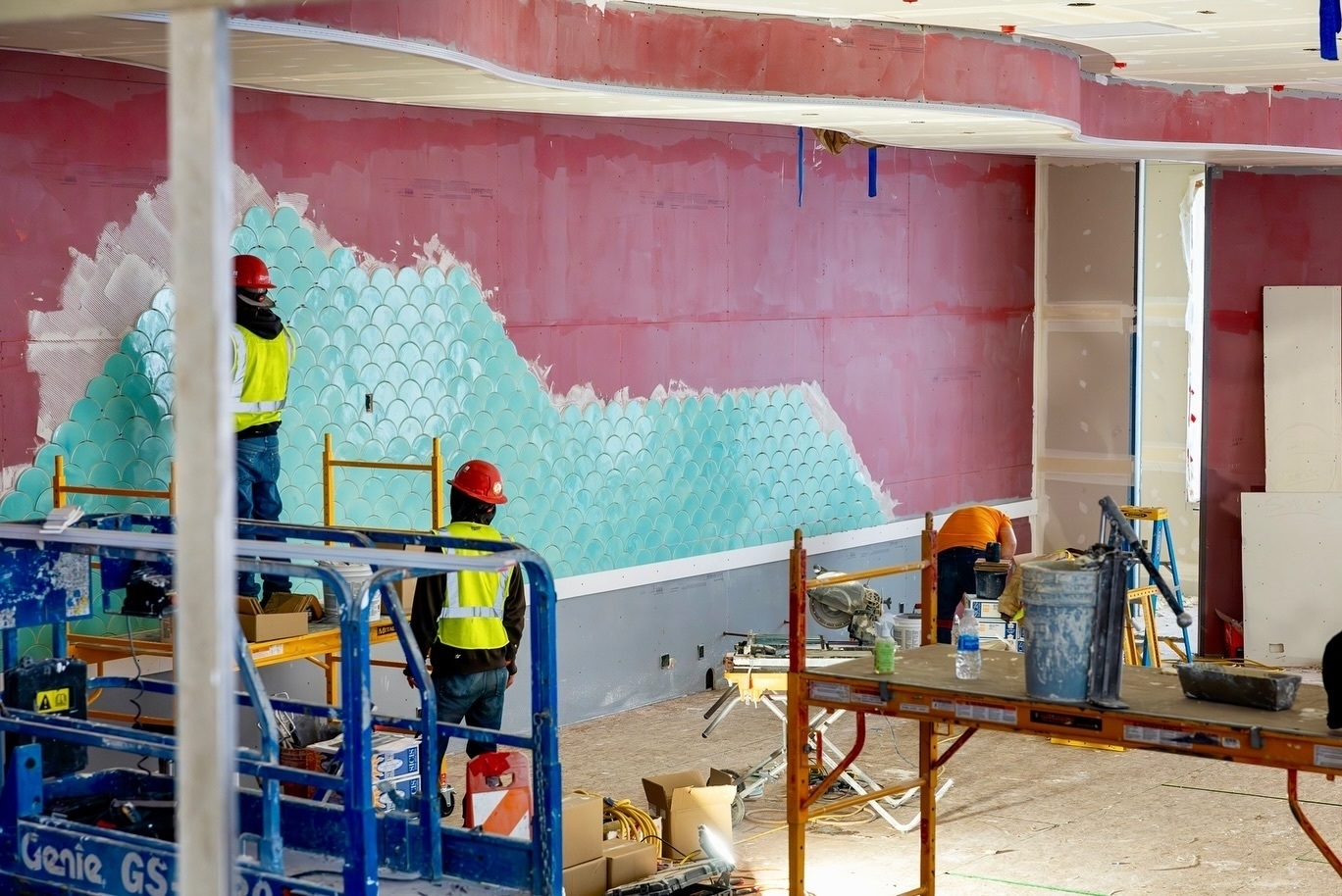 A room under construction features workers installing blue scallop-pattern tiles on a wall.