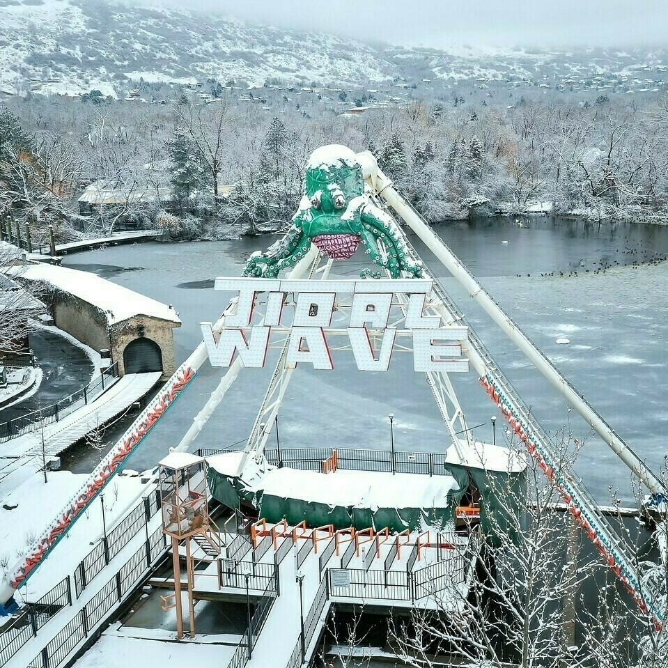 A picture of Tidal Wave in the winter. The area is covered in snow. Park of the lagoon behind the ride is covered in ice.