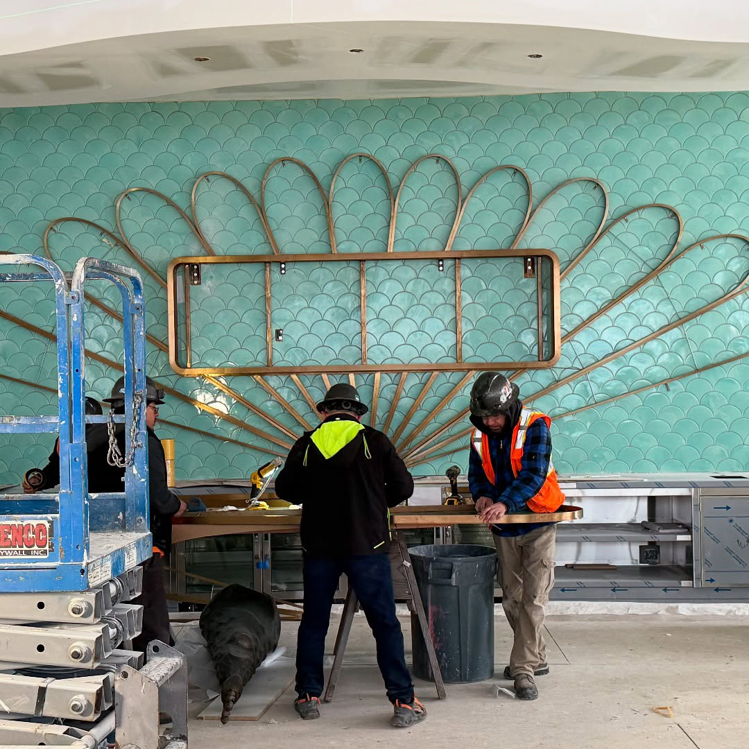 Workers in construction gear are assembling a decorative wall structure with a scallop pattern and brass elements in a room under renovation.