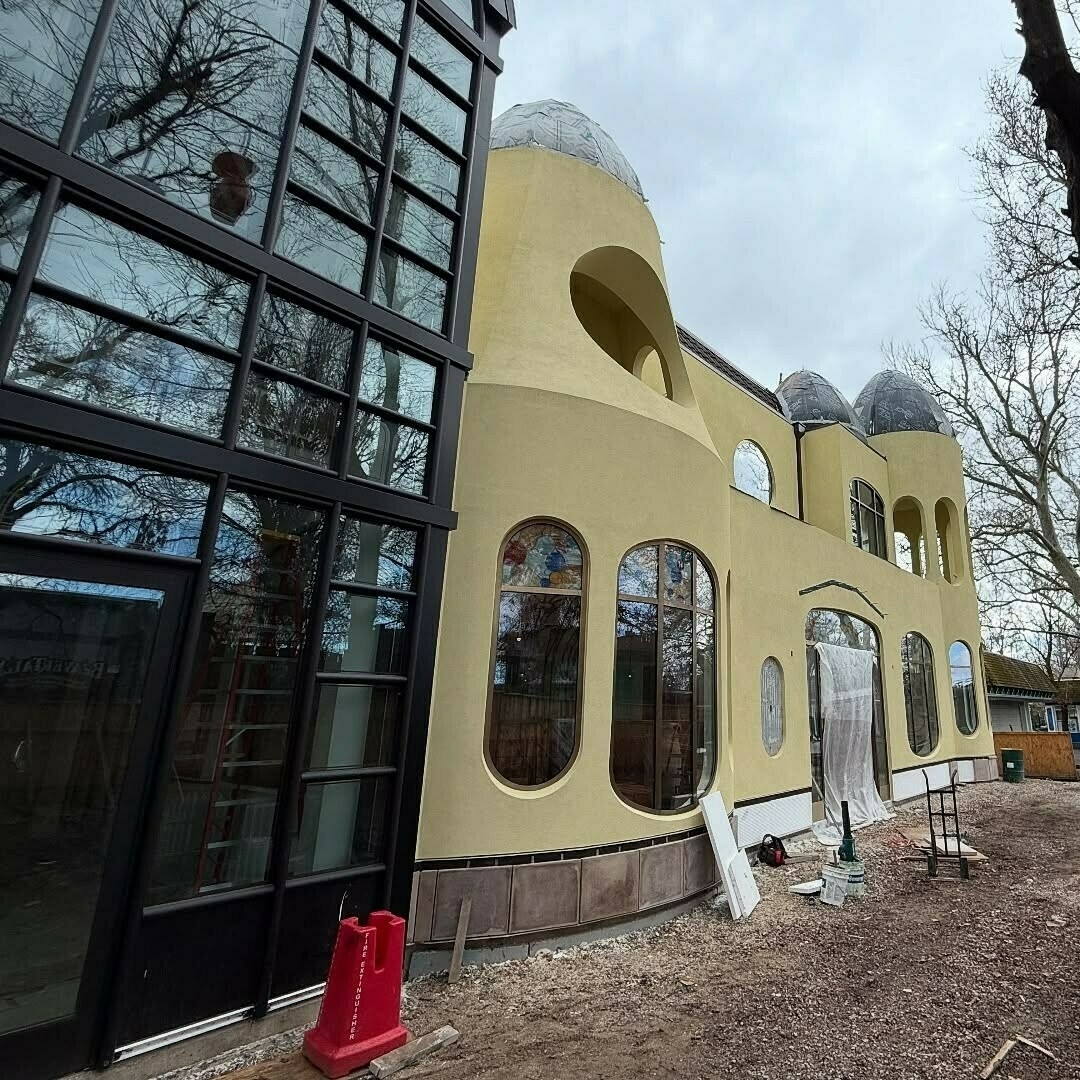 A unique building with rounded turret-like structures and large, arched windows is under construction, surrounded by trees and construction materials.