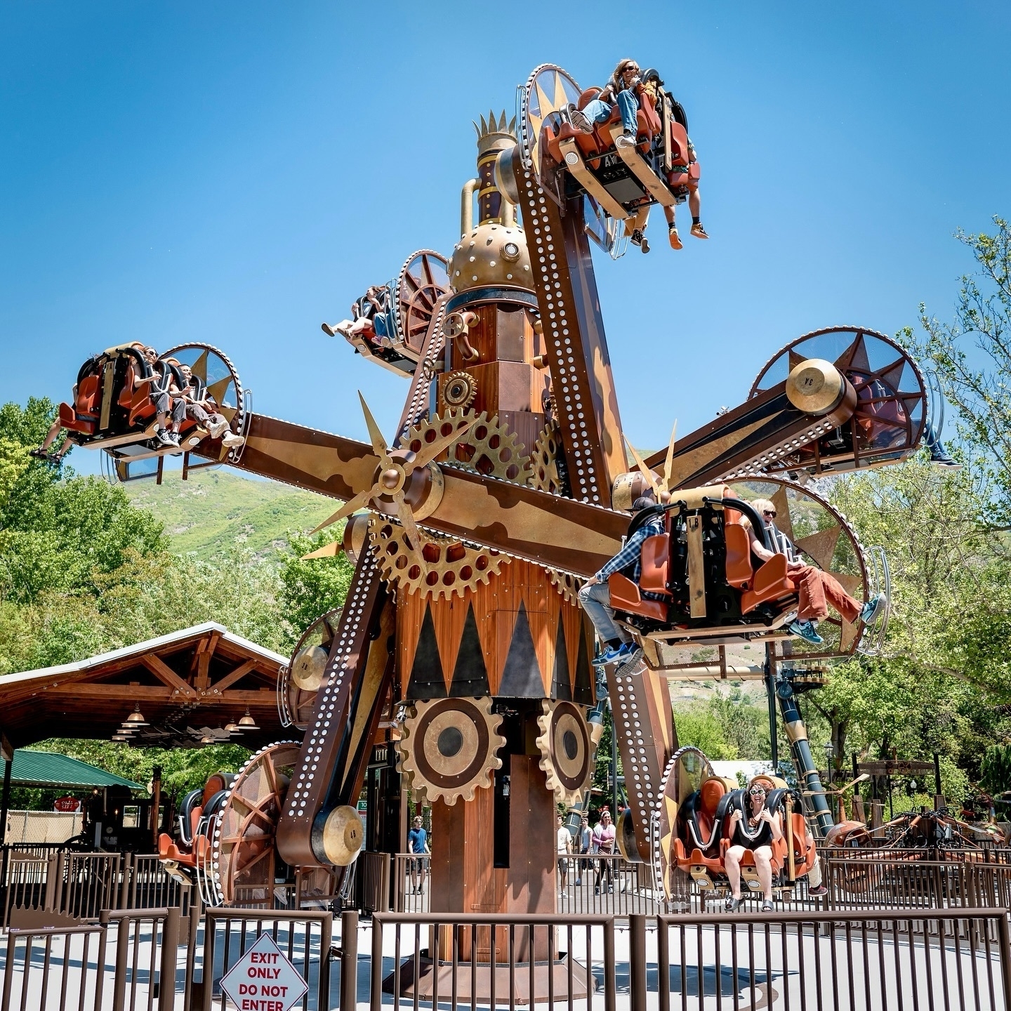 A steampunk-themed amusement ride with rotating arms and passenger seats is set against a backdrop of greenery and a clear blue sky.