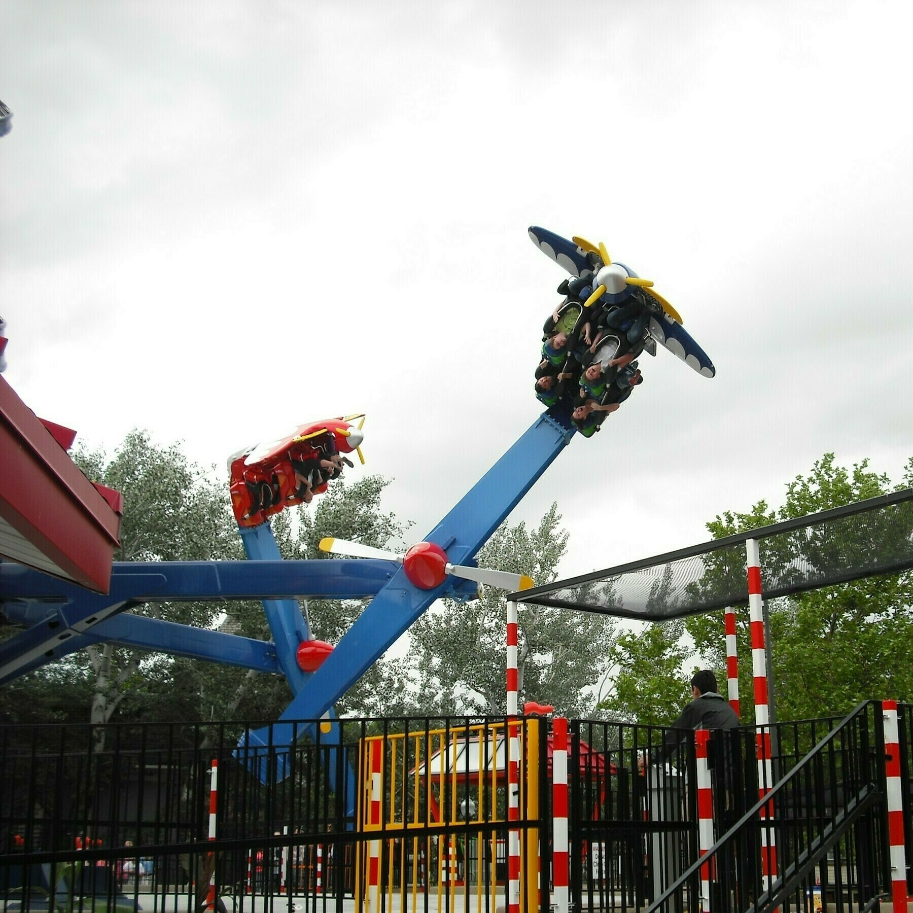 Photo of Lagoon's Air Race ride. Image is taken in early spring with cloud cover creating a white looking background. Two of the ride carriers are in the air with people going upside down. An employee can be seen in the foreground standing at an operation panel.