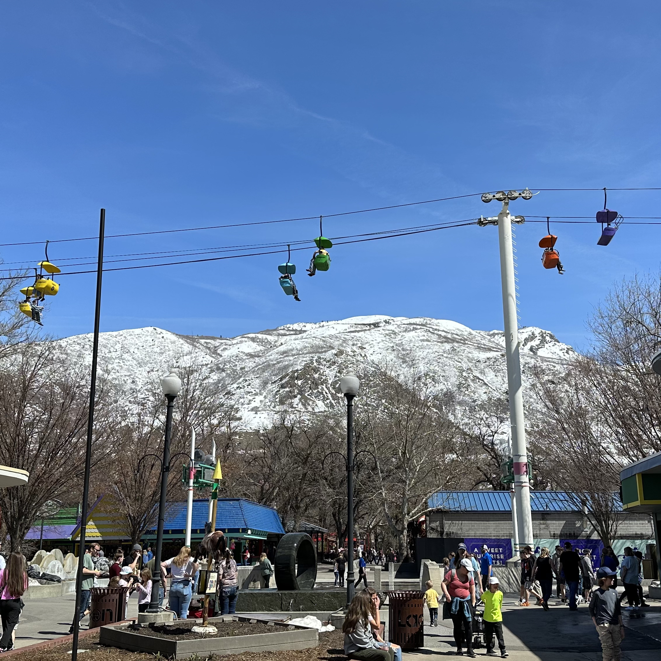 Sky Ride and Interactive Fountain area at Lagoon Amusement Park. The picture is taken from the entrance looking into the park. Mountains with snow in the background. People wandering about.