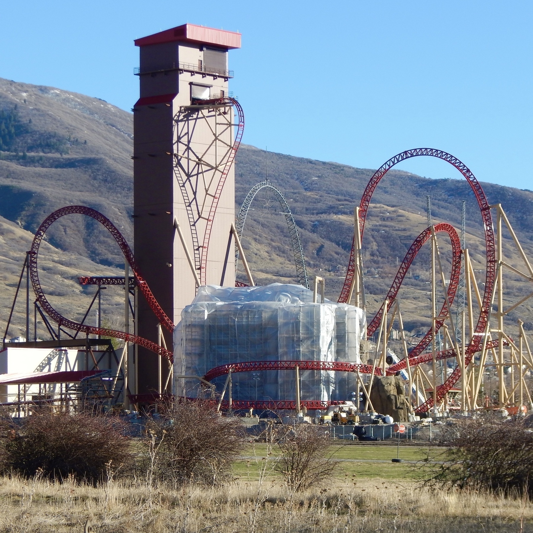 Cannibal under construction with looping tracks is set against a backdrop of hills and a tall tower structure. The large rock structure that will eventually be a waterfall is wrapped in plastic.