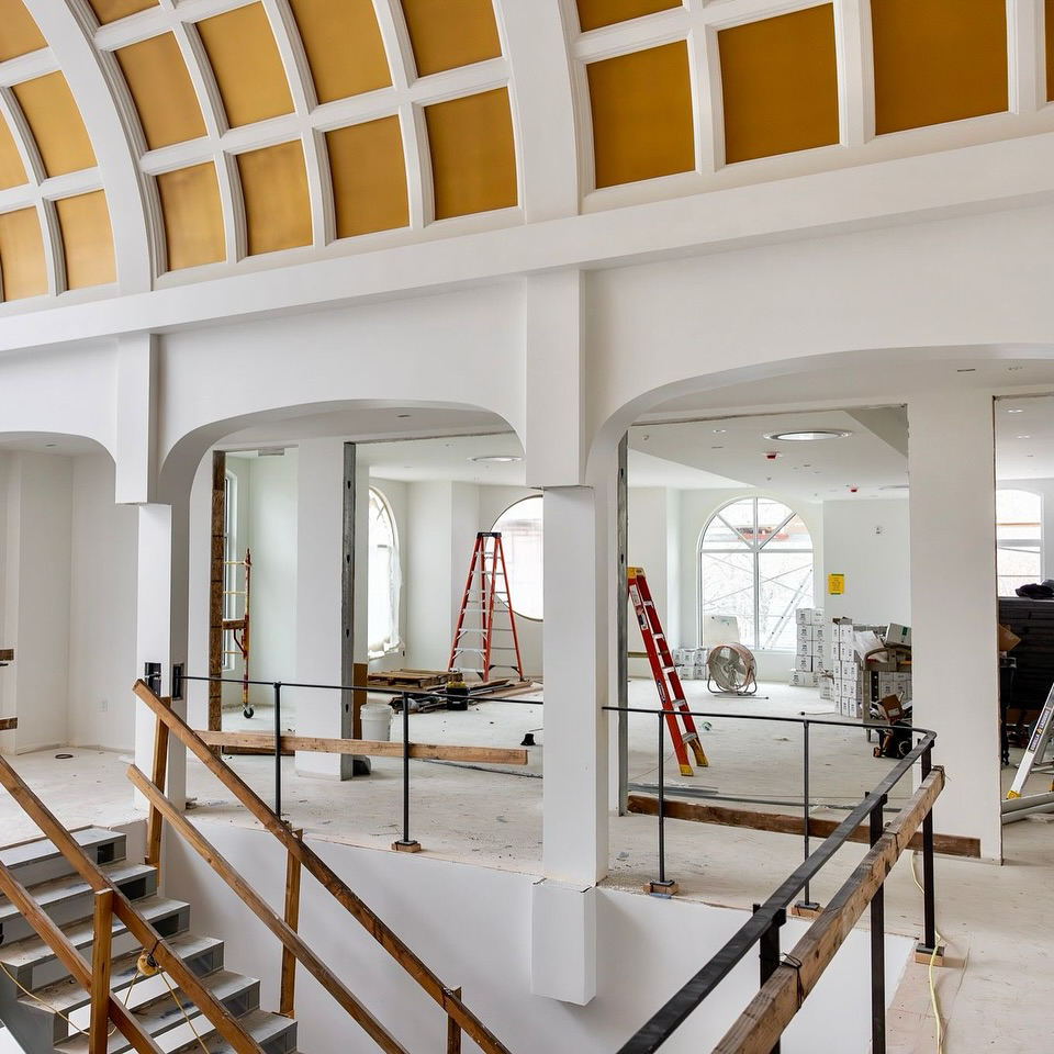 A spacious, modern interior under construction features a high, curved ceiling with yellow panels, arched windows, and scaffolding.