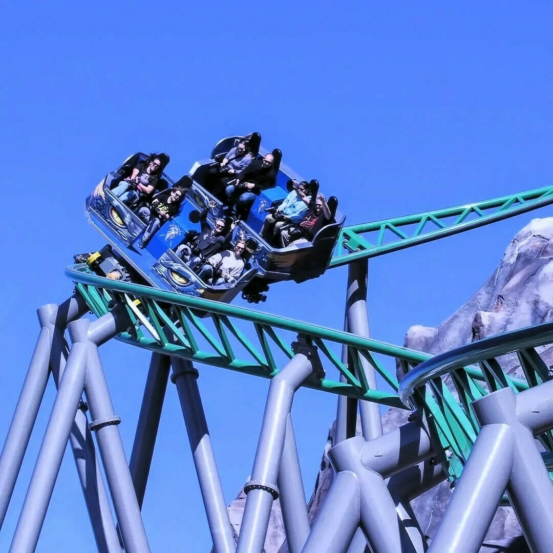 The Primordial roller coaster at Lagoon Amusement Park, featuring a green track with multiple support beams. The train of cars is filled with passengers navigating a turn, with the cars tilted to the side. The background includes a clear blue sky and part of a rocky structure, adding to the thrilling experience.