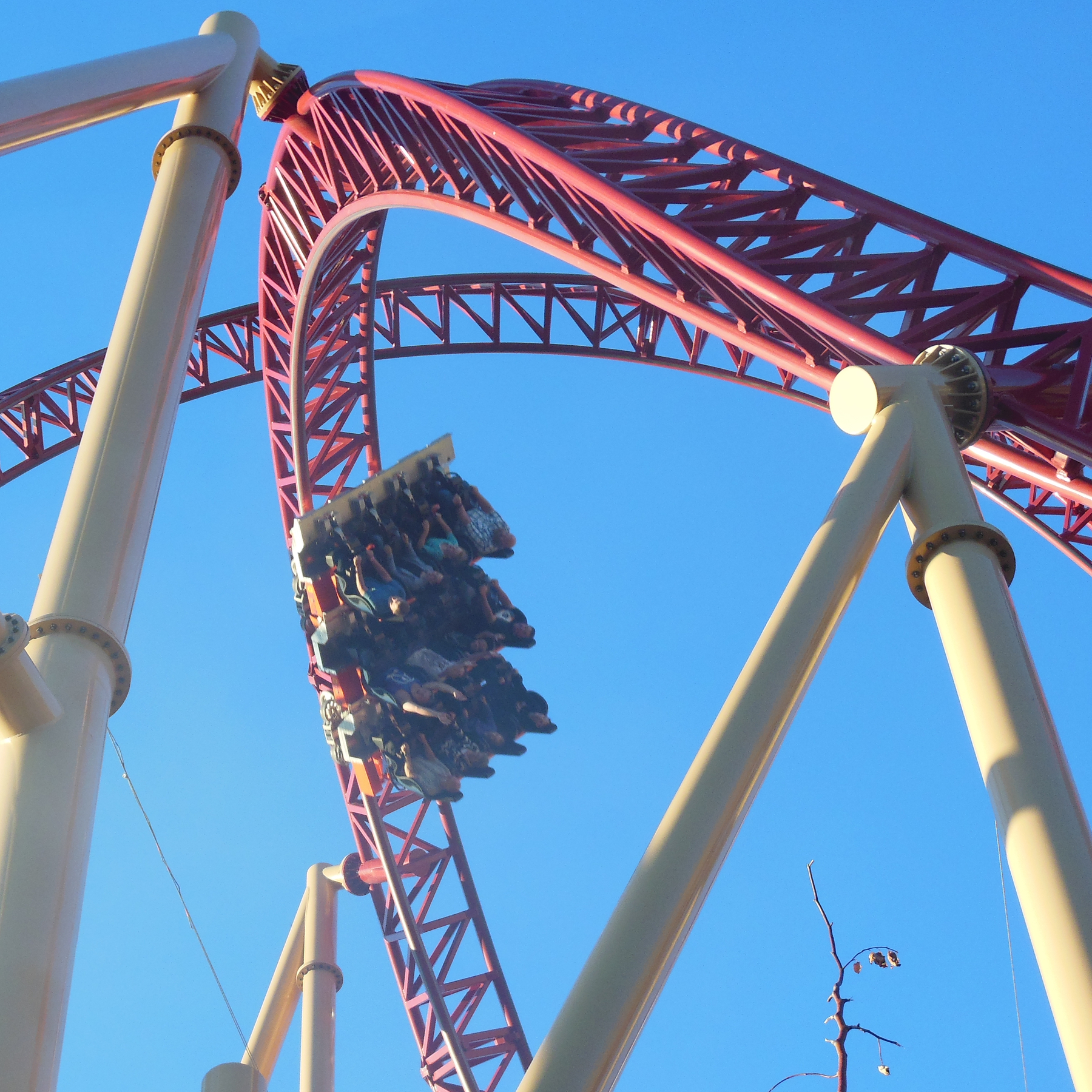A roller coaster with red tracks carries passengers upside down in a loop against a clear blue sky.