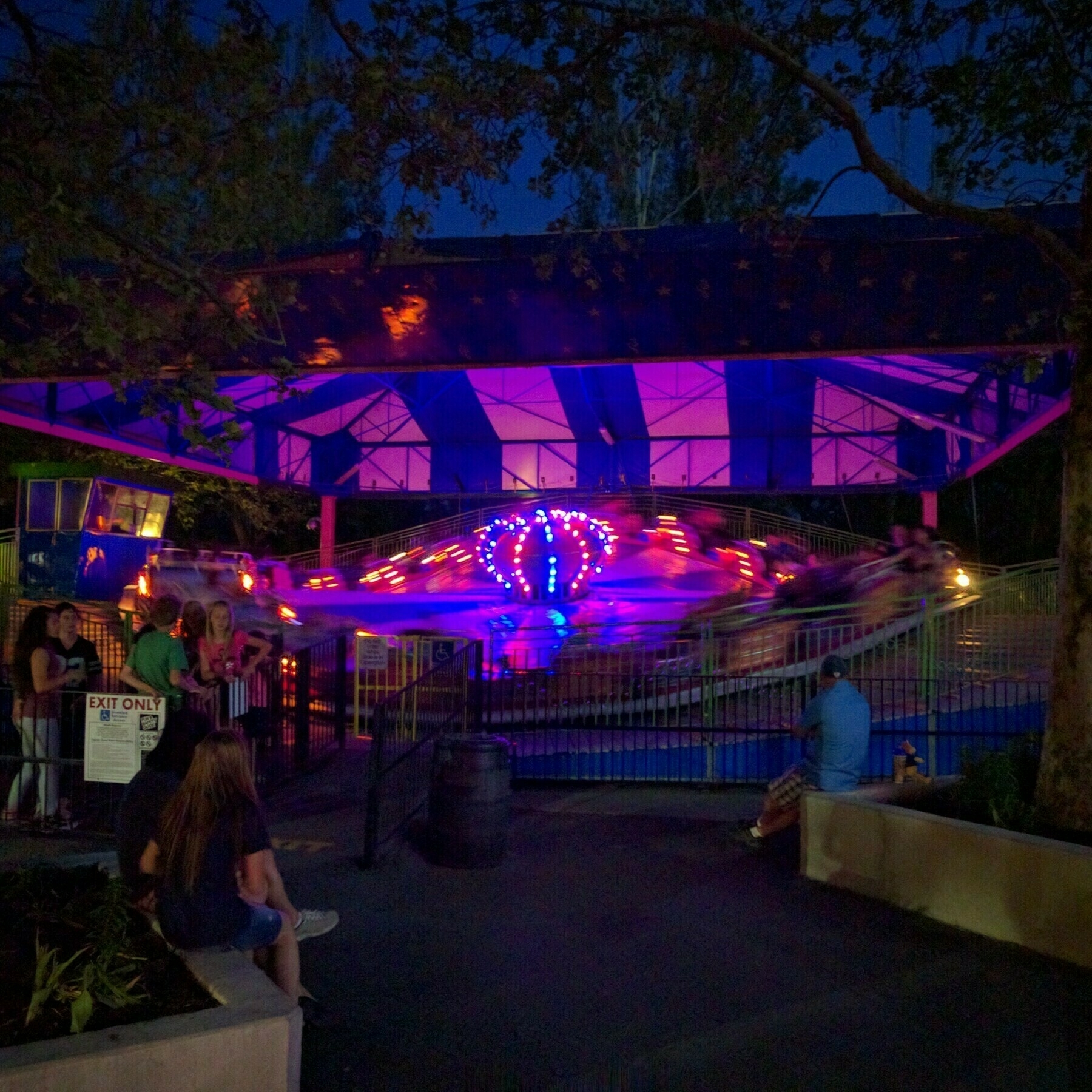 A picture of Musik Express at Lagoon Amusement Park. The image is taken at night with the ride in motion. The lights on the ride are on, and you can see some people waiting to ride as well has waiting around the ride.