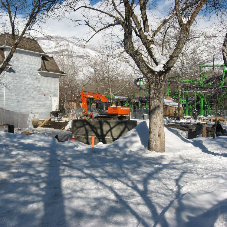 A construction site with an orange excavator and snow-covered ground. On the left in the background is the Opera House building. The Bat roller coaster is in the background on the right.