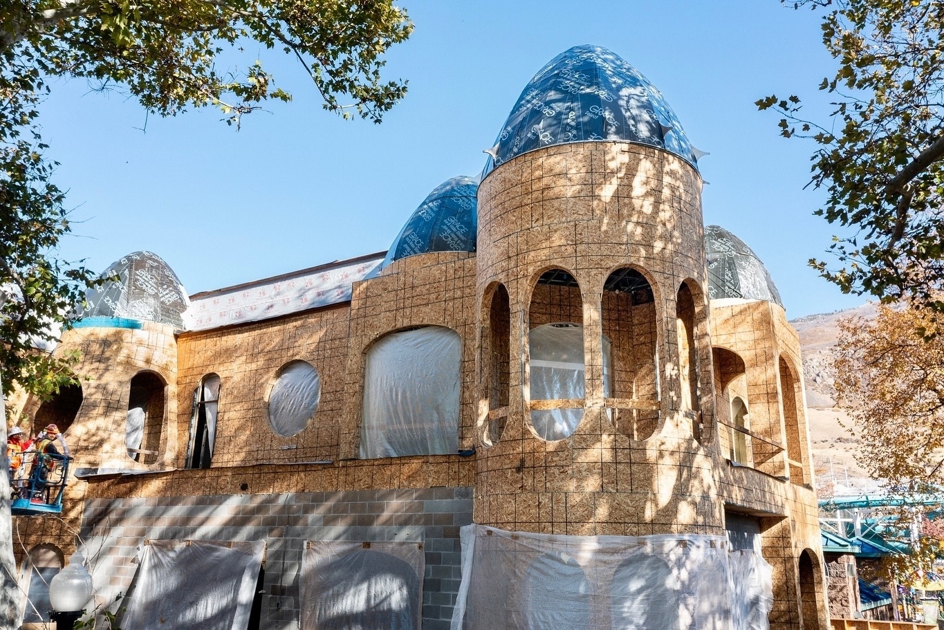 A building under construction features domed blue roofs and arched windows, surrounded by trees.