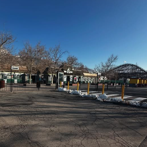 A picture taken at an angle showing the front gate of Lagoon as well as the lift hill of Roller Coaster. The sky is blue. You can see some of the parking lot with some snow piled up by the walkway.