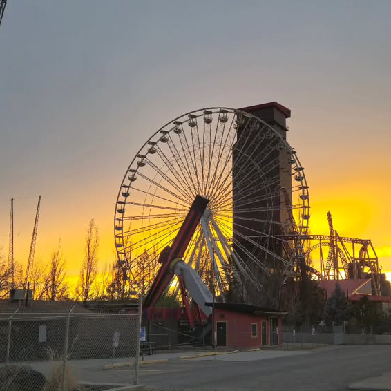 A Ferris wheel is silhouetted against a vibrant sunset sky, with surrounding amusement park structures visible in the foreground.