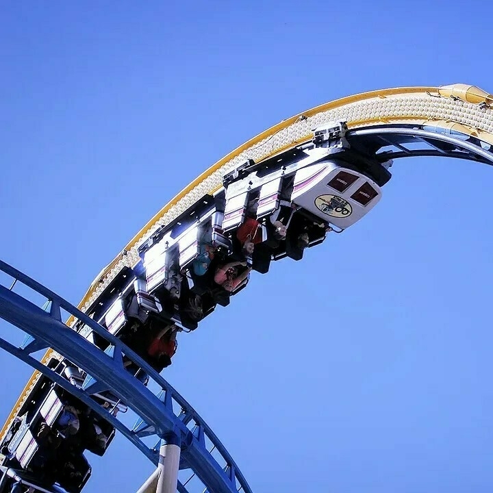 An image of the top of a loop on Fire Dragon at Lagoon. The train is at the top of the loop. People can be seen riding it. A blue curved track can be seen in front of the train.