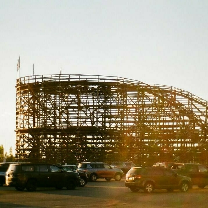 An image looking at the west turn of Roller Coaster from the parking lot taken just before sunset. Cars can be seen in front of the coaster. There is a little bit of sun glare on the image.