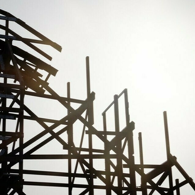 The image shows some of the beams being replaced on a wooden roller coaster. The image is captured from the ground with sunlight in the background that helps highlight the structure's details.