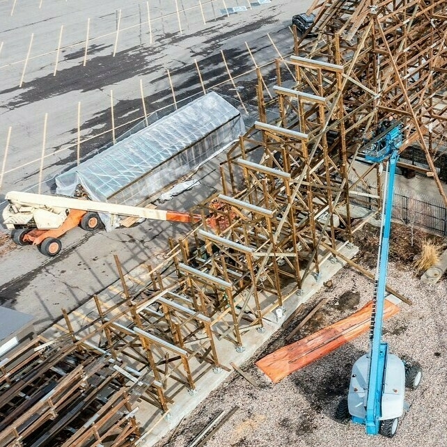 The image portrays a construction site featuring a wooden roller coaster. Two boom lifts are visible; the one on the left is orange and white, while the one on the right is blue. These lifts are being used to assist workers in accessing higher parts of the framework. The structure is composed of numerous vertical and horizontal wooden beams, showcasing the complexity and scale of the maintenance process. In the background, there's a covered structure, possibly used for storage or material protection. The ground is a mix of concrete and gravel, with various construction materials and tools scattered around, emphasizing the active and detailed nature of the site.