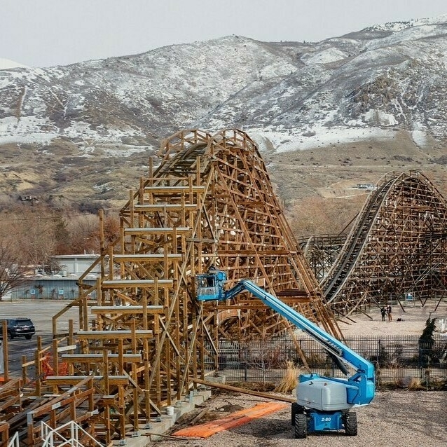 The image shows a wooden roller coaster under maintenance. A blue cherry picker lift is positioned next to the roller coaster, with a worker in the basket performing tasks on the structure. The roller coaster features a series of wooden supports and tracks, with a backdrop of snow-covered mountains and a partly cloudy sky. The scene captures the intricate process of maintaining a large amusement park ride, highlighting the scale and complexity of the structure against a scenic mountainous background.