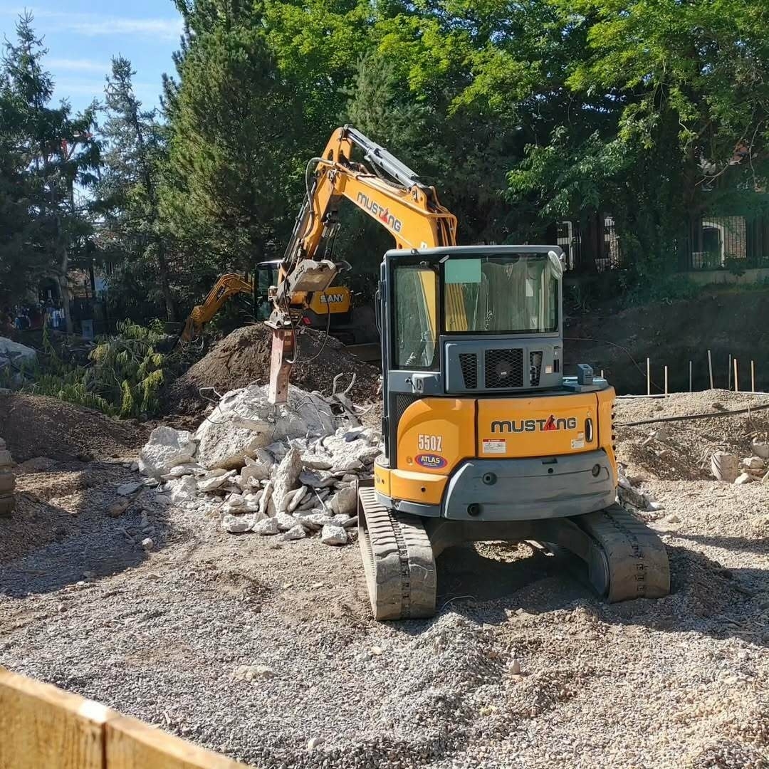 A construction site features an excavator demolishing a pile of concrete debris surrounded by trees and loose soil.